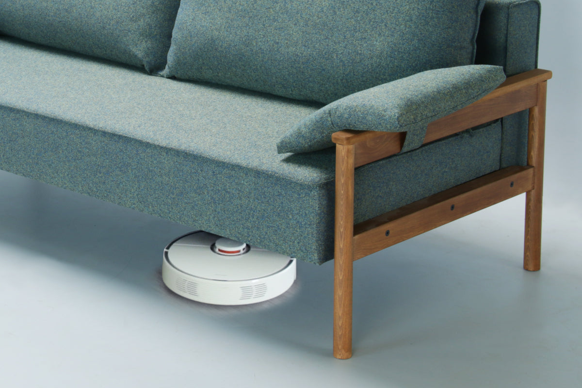 Robotic vacuum cleaner cleaning under a green Alpine sofa bed with upholstered armrest on a white background.