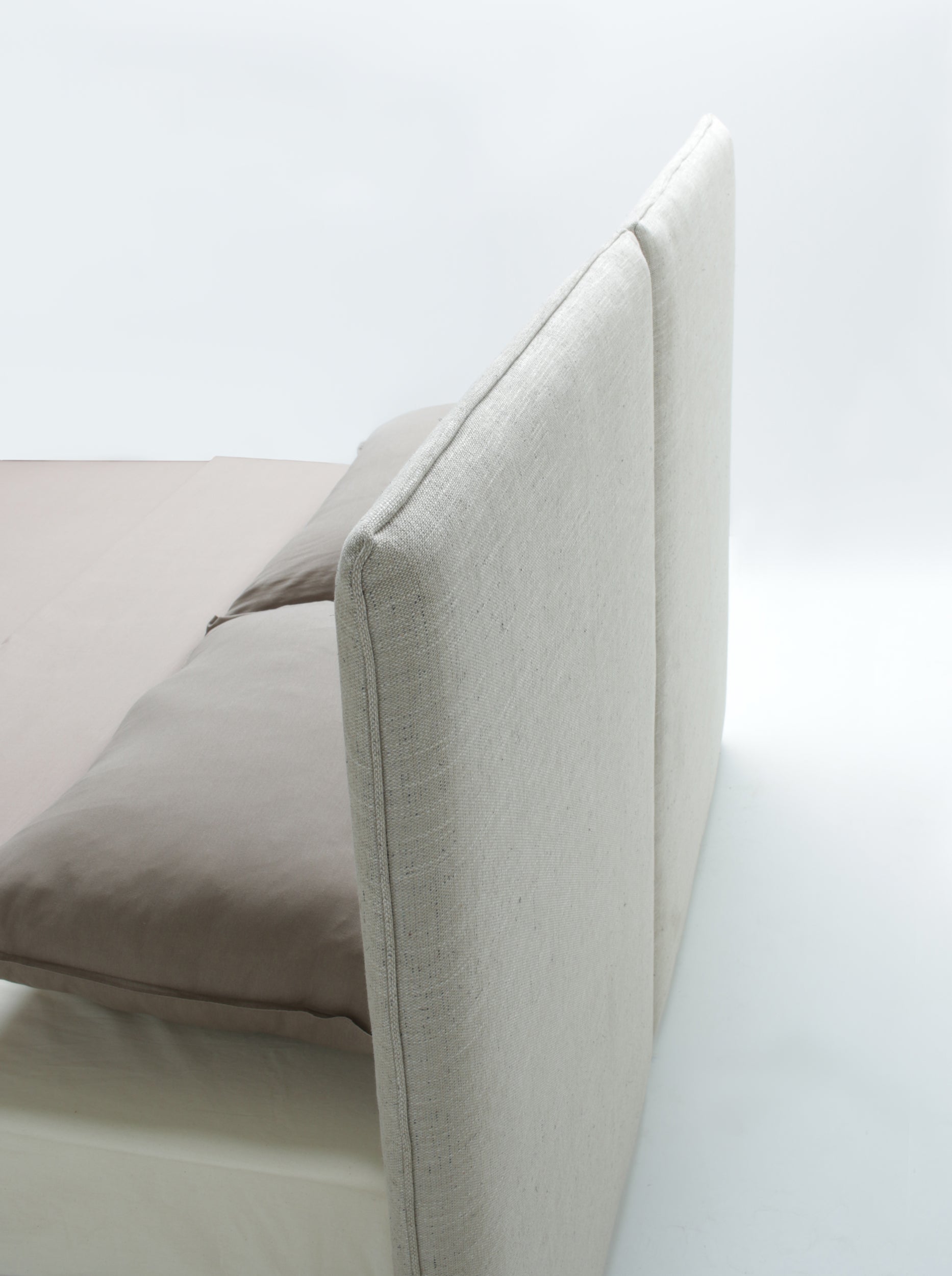Close-up of a bed frame with a tall upholstered headboard in a light beige fabric, with a taupe pillow and sheet on a white studio background.