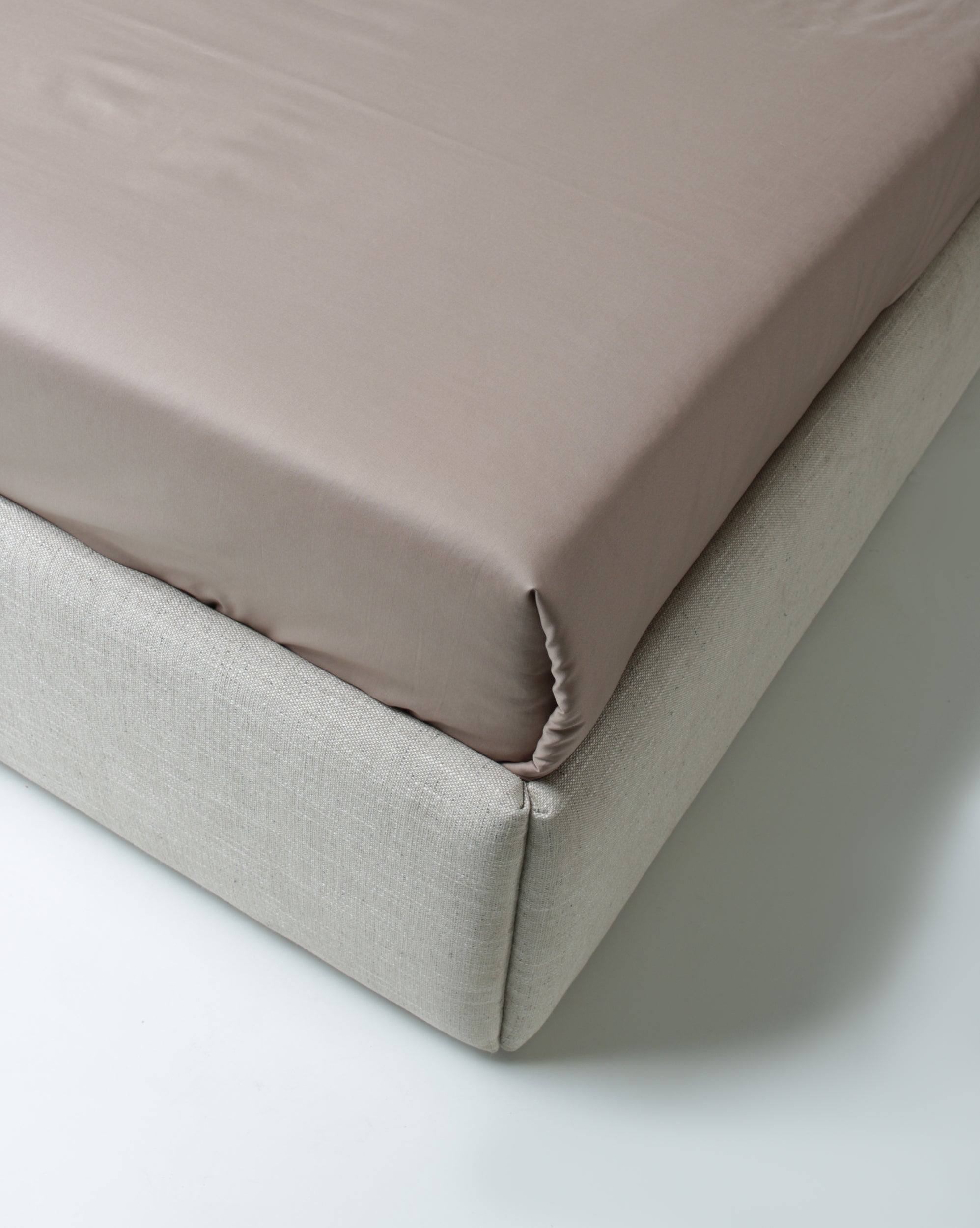 A close-up studio shot of a fitted sheet in a muted dusty pink color, pulled tightly over the corner of a light beige upholstered bed frame.
