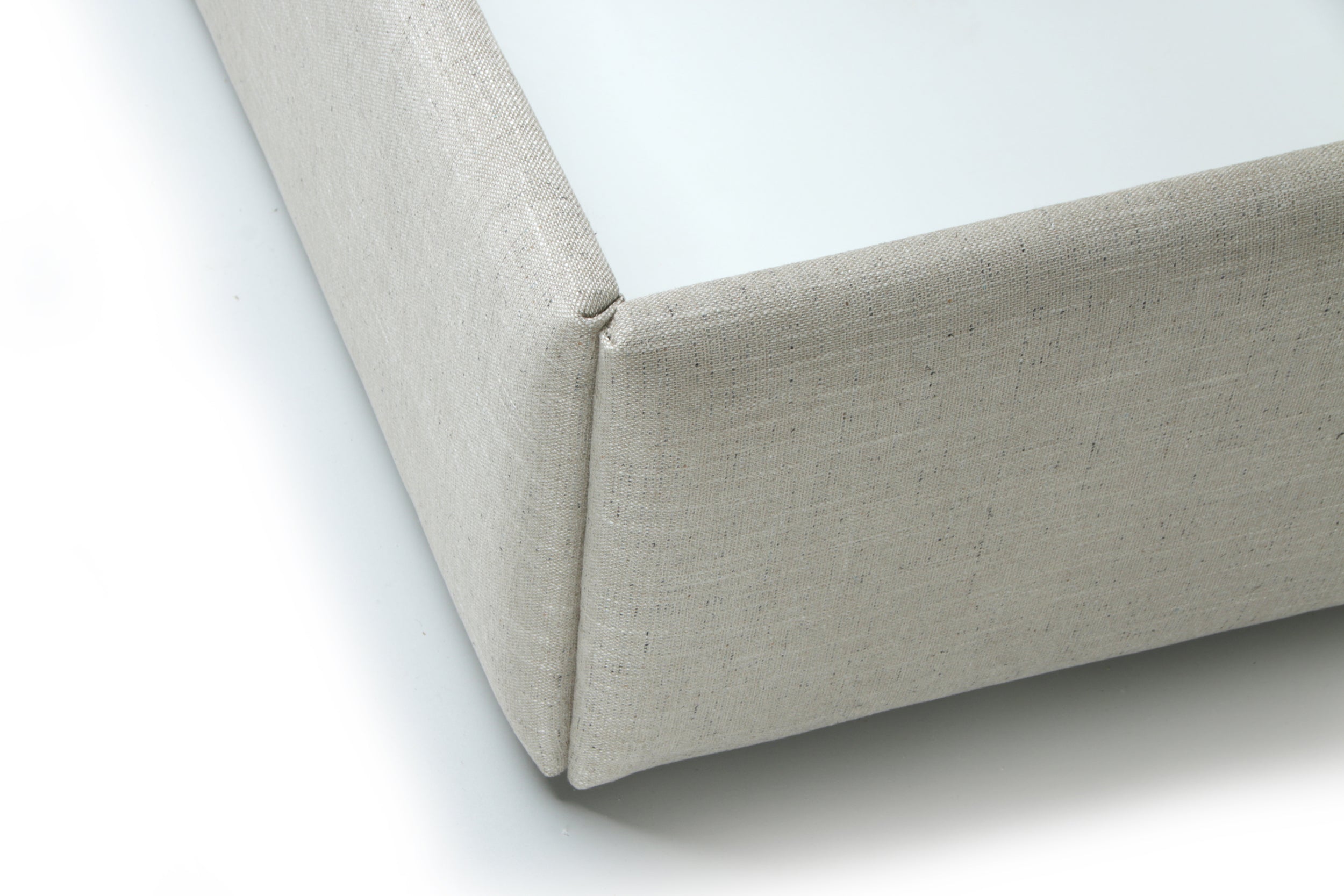 Close-up of a light beige upholstered bed corner, showing the fabric texture and folded edge detail on a white background.