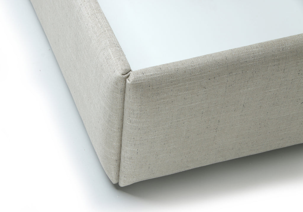 Close-up of a light beige upholstered corner of a boxy furniture piece, showing the fabric texture and clean white surface underneath on a white background.