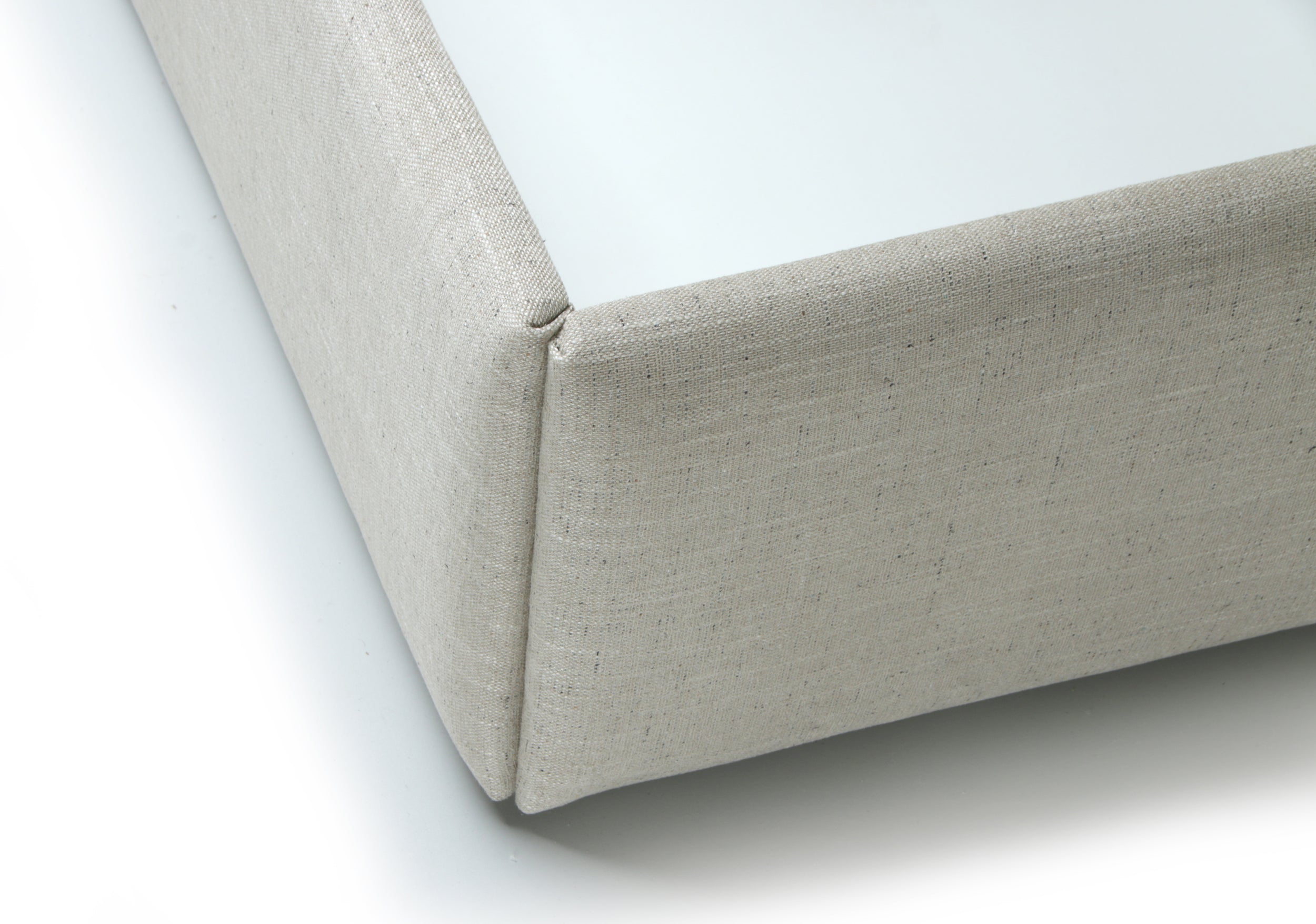 Close-up of a light beige upholstered corner of a boxy furniture piece, showing the fabric texture and clean white surface underneath on a white background.