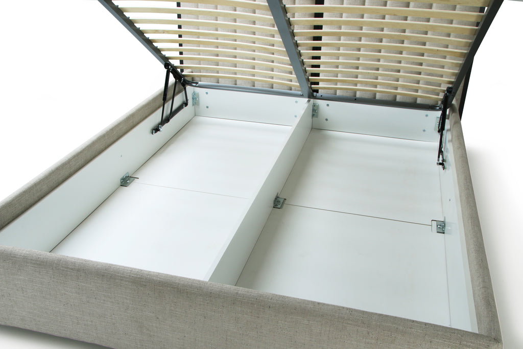 A close-up of a bed frame with the lift-up slatted lid open, showing the light gray upholstered sides, white storage compartments, and beige slats inside.
