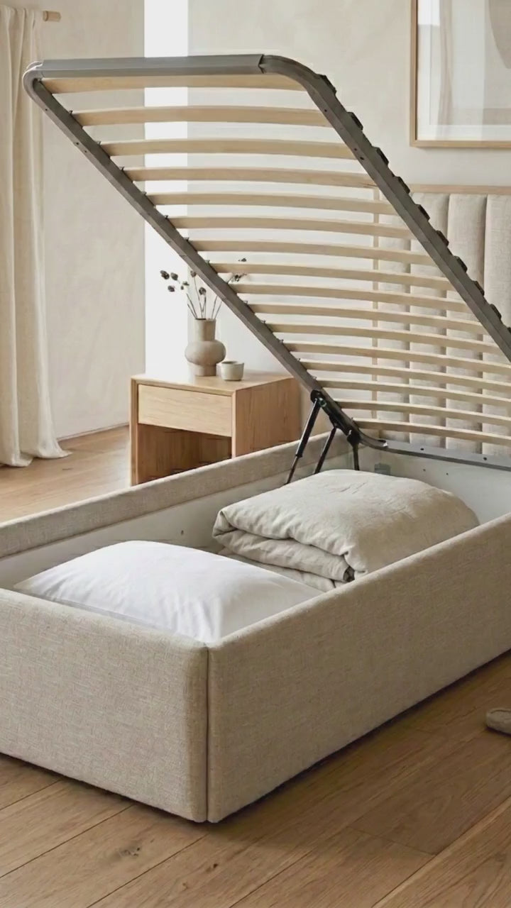 Beige upholstered bed with open chest in a room with a framed picture on the wall and upholstered  headboard.